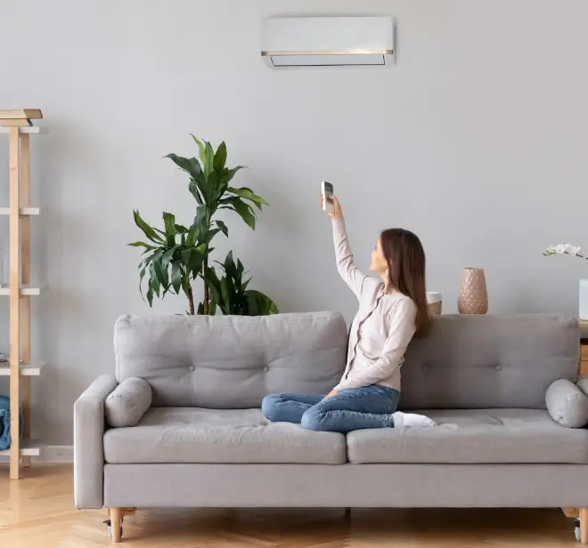 Woman sitting on a gray sofa using a remote control to adjust the wall-mounted air conditioning unit, with a plant and decorative items in the background, illustrating indoor comfort and HVAC control.