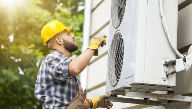 Technician in a yellow hard hat and gloves inspecting an outdoor air conditioning unit, ensuring system efficiency and safety during maintenance.