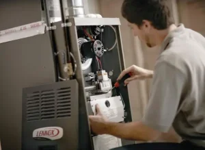 A technician in a tan shirt uses a screwdriver to repair the internal components of an open Lennox gas furnace.
