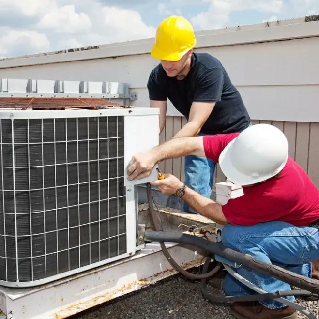 Two HVAC technicians servicing an air conditioning unit on a rooftop, wearing hard hats, engaged in repair work, showcasing expertise in HVAC maintenance.